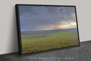 A black framed canvas depicting a scenic landscape with rolling hills, a field of grass, and a dramatic sky with clouds and a sunset.
