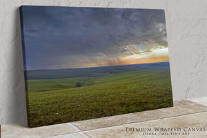 The image is a landscape painting depicting a vast, rolling green field with a dramatic sky filled with clouds and a vibrant sunset.