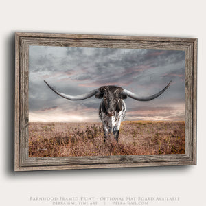 A framed print depicting a large horned bull standing in a field with a cloudy sky in the background.