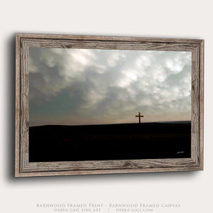 A framed art print featuring a dramatic landscape with a lone wooden cross on a dark horizon beneath a cloudy sky, presented in a rustic barnwood frame against a light background.