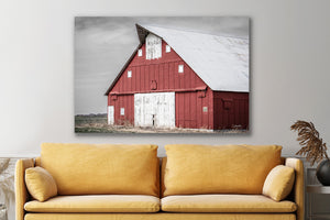 A large rectangular wall art print showing a red barn with a white roof, displayed above a mustard yellow sofa in a modern living room setting.