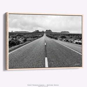 A framed black and white landscape photograph of a desert road leading toward buttes, shown as a floating canvas print with a natural maple frame against a light gray background.