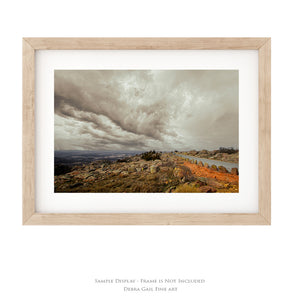 The image is a framed photograph depicting a rocky landscape with a body of water in the background, under a cloudy sky.