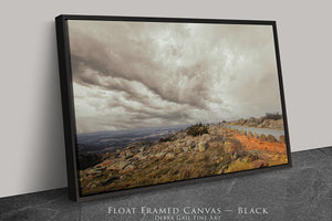 A black framed canvas depicting a scenic landscape with a rocky hillside, a body of water, and a cloudy sky.