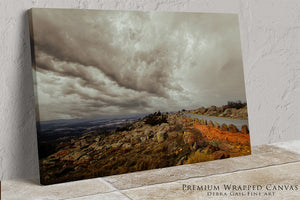 The image is a landscape photograph featuring a rocky hillside with a winding road, set against a cloudy sky.