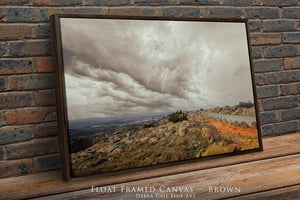 A framed landscape photograph depicting a rocky hillside with a winding road and a cloudy sky in the background.