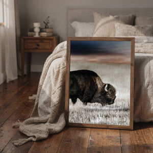 A framed art print of a bison in a grassy field, leaning against a bed in a cozy, neutral-toned bedroom.