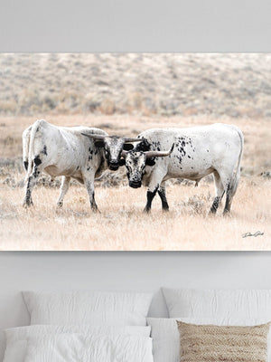 A canvas print of two longhorn cattle interacting in a field, mounted on a wall above a bed in a room with a neutral color scheme.