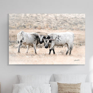 A canvas print of two longhorn cattle interacting in a field, mounted on a wall above a bed in a room with a neutral color scheme.